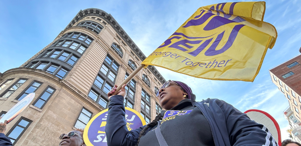 From a heroic low angle, we look up at a Black woman marching in a purple SEIU BJ shirt. The yellow SEIU flag she carries is billowing in the wind, mirroring the curve of the building behind her, against the blue sky. We can see the corners of other picket signs--she is part of a dense crowd.