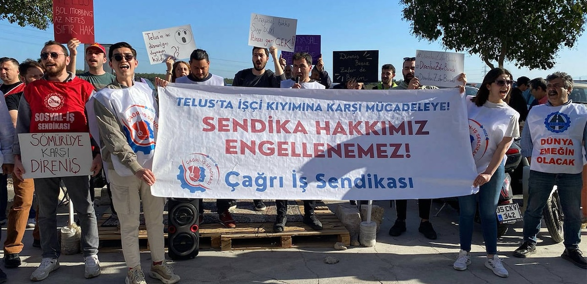 A group of 30 men and women with signs in Turkish stand outside behind a banner