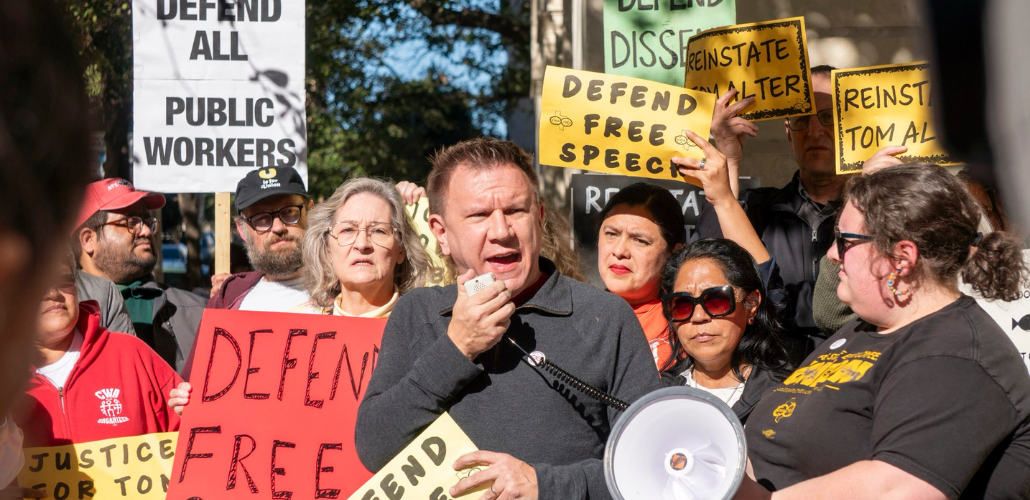Tom Alter, a white man with a short haircut, squints into the sun and speaks into a megaphone, He is surrounded by a tightly packed crowd, diverse in race and gender, holding signs with slogans like "Defend Free Speech," "Defend All Public Workers," and "Reinstate Tom Alter."