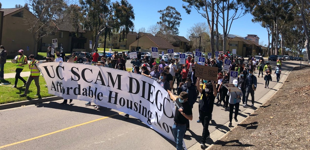 A group marches down the street behind a banner reading "UC Scam Diego: Affordable Housing Now."