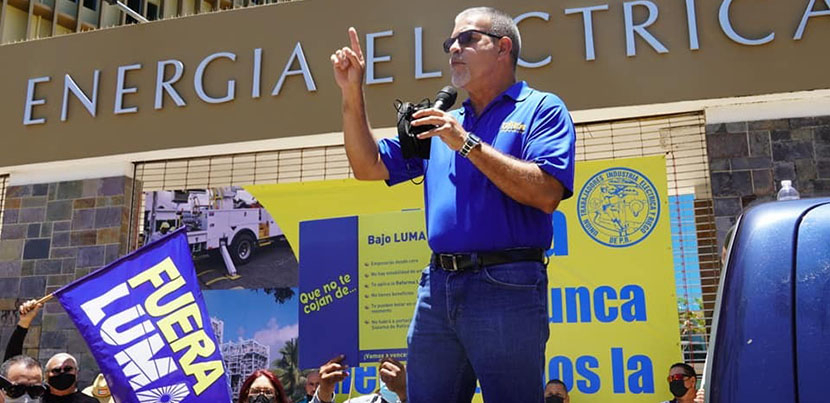 Man in blue shirt with right arm raised and left arm holding microphone addressing crowd, Fuera LUMA flag visible in foreground, building of Puerto Rico Electric Power Authority (PREPA)