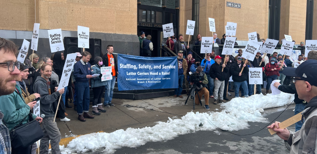 Postal workers rally outside a post office. There is some snow on the ground. A big blue banner says "Staffing, Safety, and Service: Letter Carriers Need a Raise! NALC Branch 9, Minneapolis." Printed picket signs say "End Mandatory Overtime," "First Class Service, First Class Pay," and "Fair Contract Now."