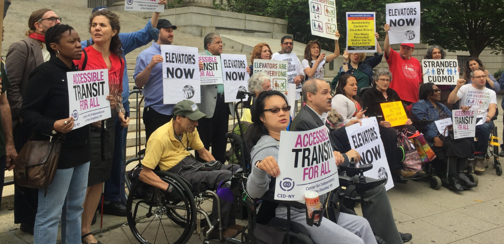 Demonstrators, some in wheelchairs and some not, stand in front of a public building. Many are holding signs: "ACCESSIBLE TRANSIT FOR ALL," "ELEVATORS NOW," "STRANDED BY CUOMO"