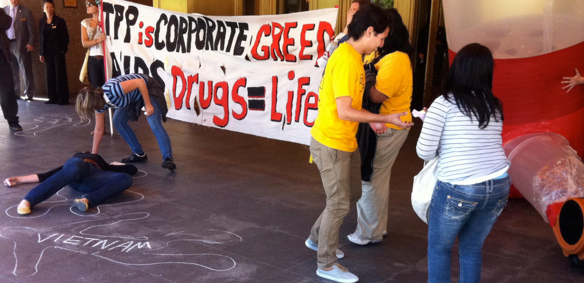 Activists draw chaik outlines on the ground in front of a hotel entrance; one outline is labeled "Vietnam." Others hold a banner: "TPP is corporate greed. AiDS drugs = life."
