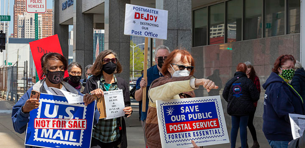 Picketers (Black and white, men and women) marching in front of a post office. Signs say "U.S. Mail Is Not for Sale," "Postmaster DeJoy Is Destroying Our Post Office," "Save Our Public Postal Service: Forever Essential." Also visible are copies of the flyer "Dump DeJoy and His 10-Year Plan."