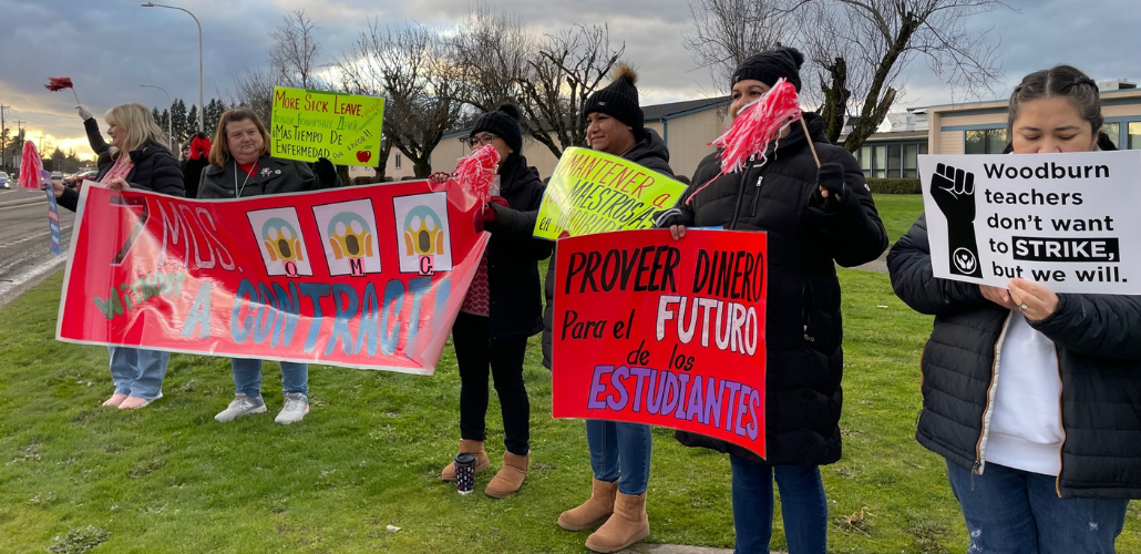 People stand on grass with signs. A red banner says "Seven months without a contract, OMG!" with stylized images of Edward Munch's painting "The Scream." Another sign says "More sick leave" in English, Spanish, and Russian. Another says, in Spanish,"Provide money for the future of students." Another says "Woodburn teachers don't want to strike, but we will." 