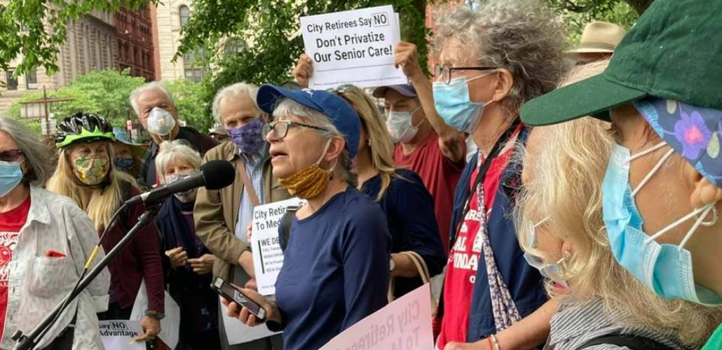 A group of older people in masks stands outside. The person in the middle is speaking into a mic. Someone behind her holds up a printed sign: "City Retirees Say NO, Don't Privatize Our Senior Care!"