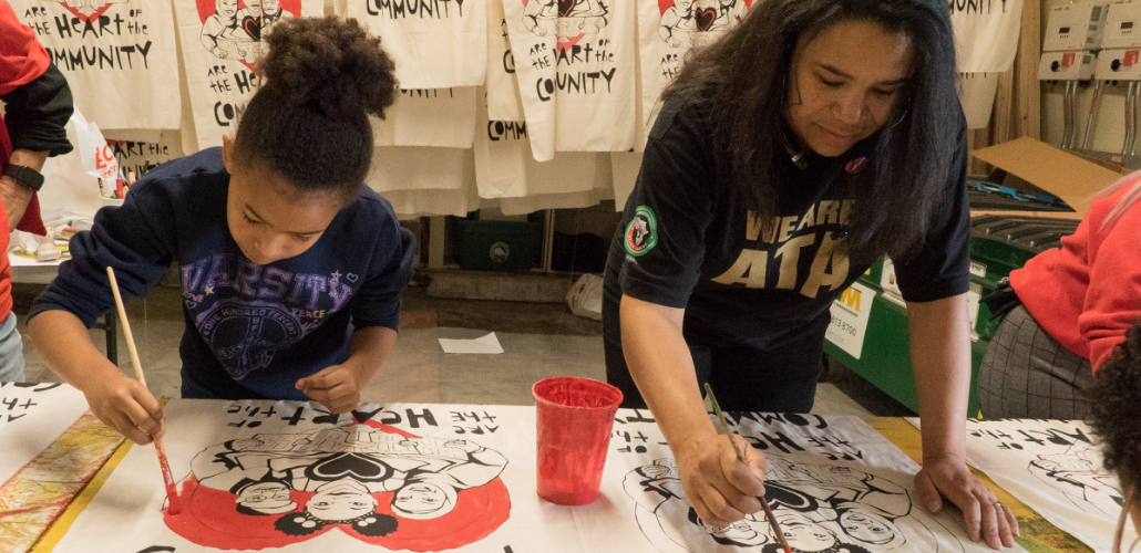 A child and an adult paint signs. Behind them are many more signs hanging already painted.