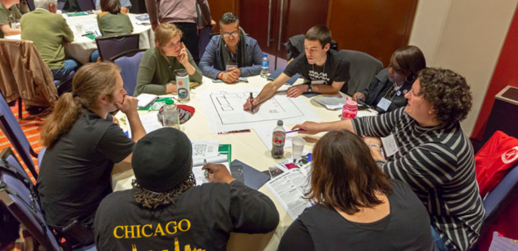 People sit around a big table, leaning in. One is drawing a workplace map on big paper, while another points something out. They're in a hotel conference room. Other table groups are visible behind them.