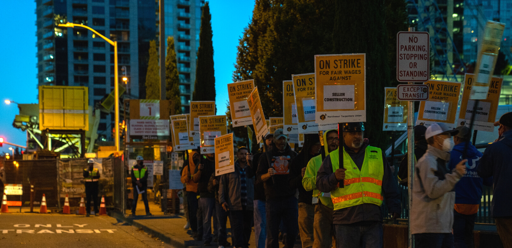 Picketers in yellow vests holding "ON STRIKE FOR FAIR WAGES AGAINST SELLER CONSTRUCTION" signs outside in dim dawn light.