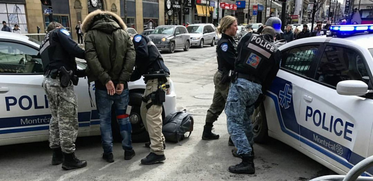 Outdoors, four police officers handcuffing two people against two police cars