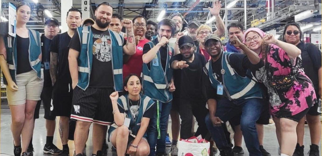 A group of workers, diverse in race and gender, poses together on the factory floor. Most appear young; one wears a prosthetic arm. A few have fists raised and most smile or wear determined expressions. Most wear shorts and some wear blue work vests.