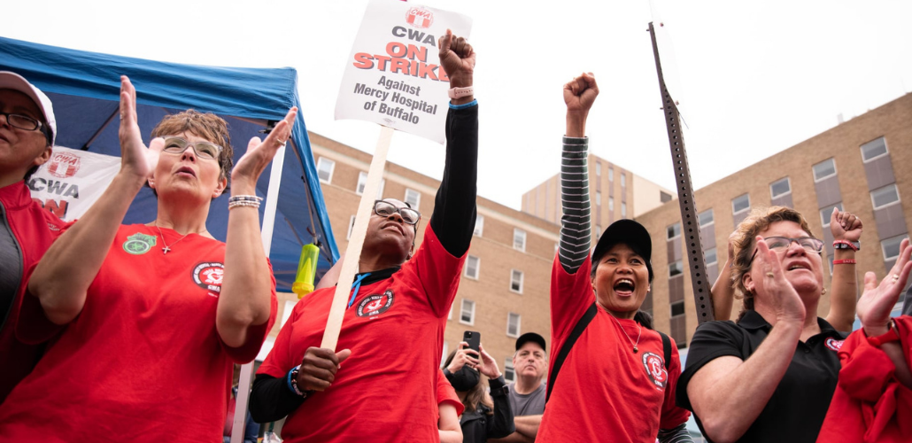 Joyful people in red shirts throw their fists in the air. One has a "CWA On Strike Against Mercy Hospital of Buffalo" picket sign. Outdoors.