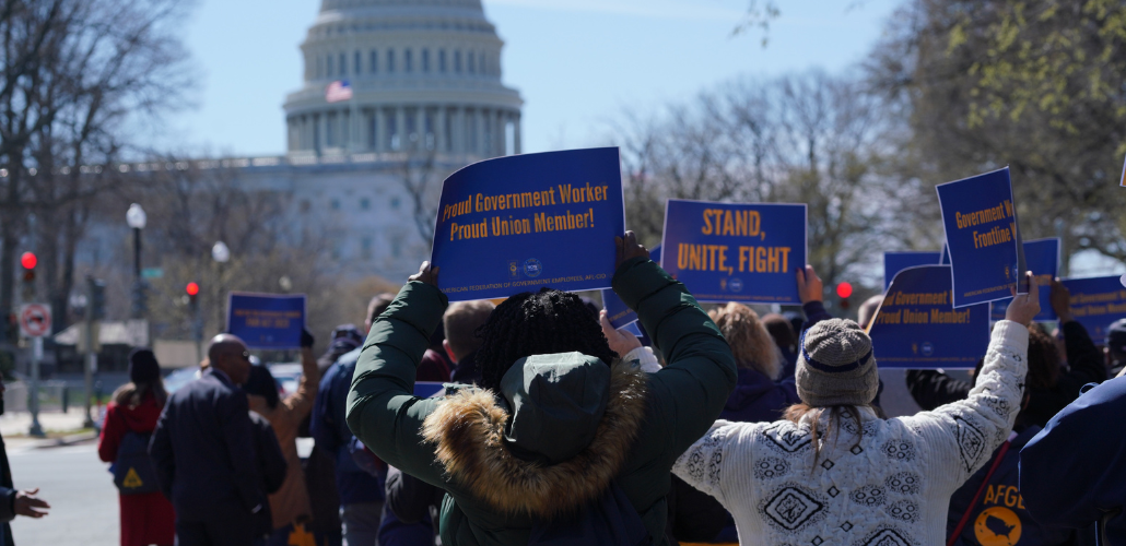Crowd of people viewed from behind, holding blue AFGE sign. One says "Proud Government Worker, Proud Union Member. Another says "Stand, Unite, Fight." In front of them is the U.S. Capitol building.