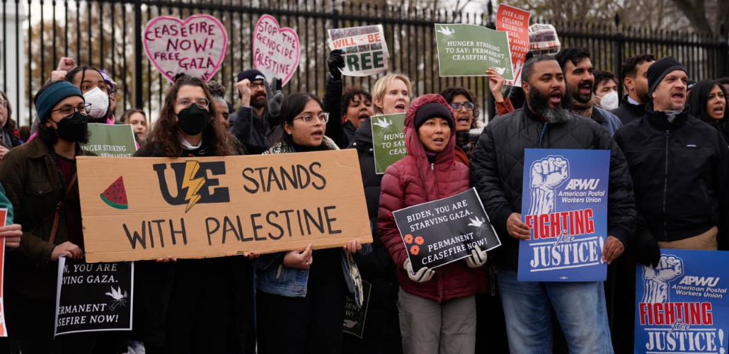 A chanting crowd stands in front of the White House fence. Three people are helping hold a large cardboard sign that says "UE stands with Palestine" with the union's lightning-bolt logo and a slice of watermelon. Other printed signs say "American Postal Workers Union: Fighting for Justice" and "Biden, you are starving Gaza. Permanent ceasefire now!"