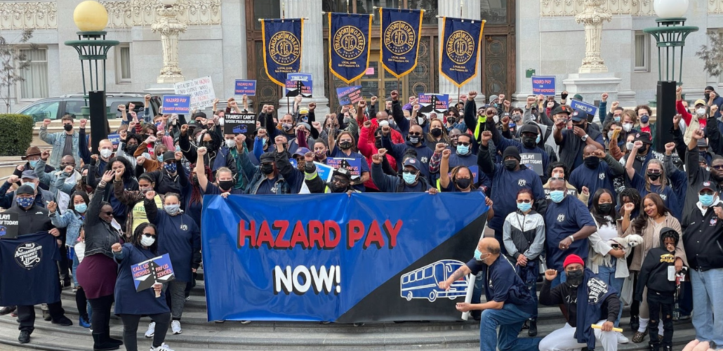 Large, multiracial, masked crowd stands on the steps of a public buliding. Many have fists in the air. Back row holds ATU banners. Front row holds a giant banner that reads "Hazard pay now!" with a line drawing of a bus.