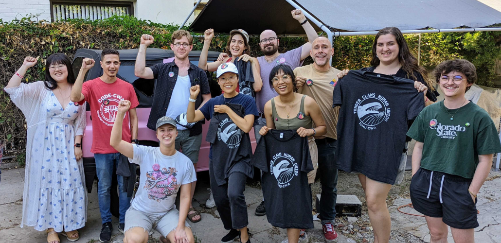Eleven smiling people pose outside. Some are holding up silkscreened T-shirts with Tender Claws union logo showing a claw. Many are raising one fist, and one is pointing at another person in the group.
