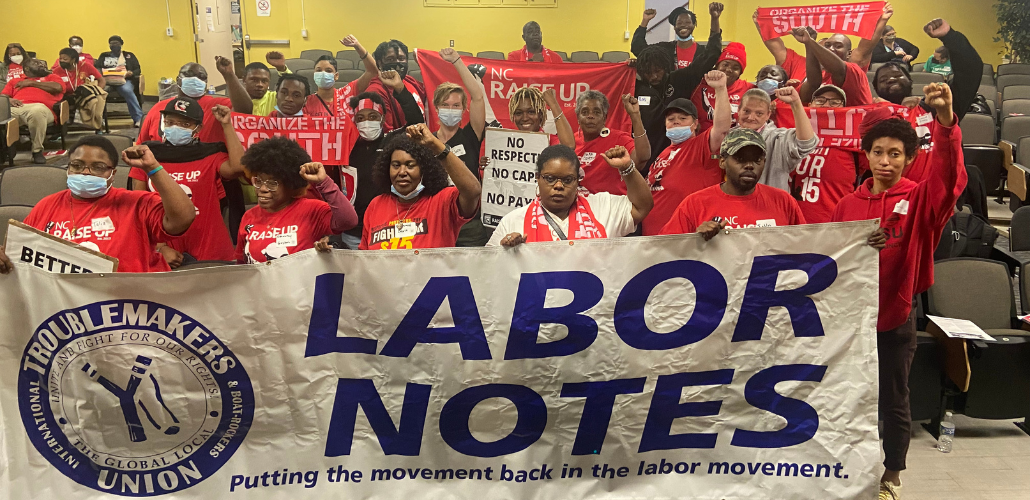 People stand indoors behind a big Labor Notes banner. Most are wearing red shirts and have their fists in the air. Several farther back are holding up smaller red "Organize the South" banners.