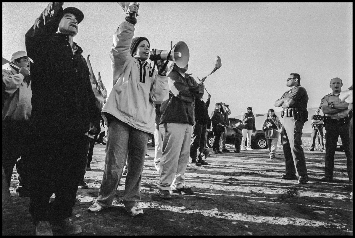 A vintage black-and-white photo shows a line of workers standing outside, one calling into a megaphone, facing down cops with folded arms. They are photographed from a low, dramatic angle.