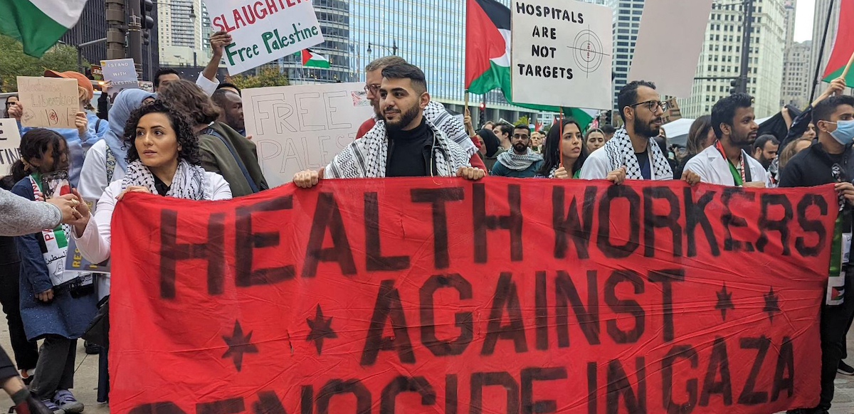 Workers stand outdoors, many wearing keffiyehs, holding a large red hand-painted banner that says "Health care workers against genocide in Gaza." Signs visible in crowd behind them say "Hospitals are not targets," "Free Palestine," and something ending in "slaughter." 