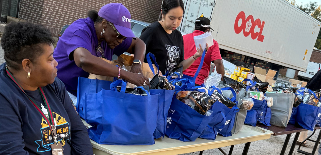 Three women work around a table with blue cloth grocery bags, outdoors. A man in a red T-shirt walks behind them. A truck is also visible.