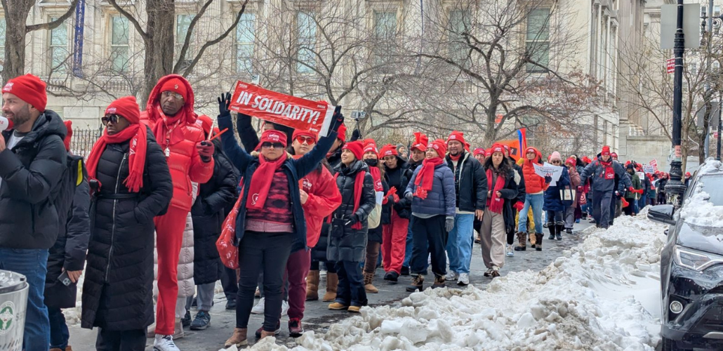 A long procession of strikers bundled up against the cold, most wearing bright red NYSNA hats or scarves, packs a sidewalk lined with piles of shoveled snow. The group is diverse in race and gender. Many are smiling. A woman in the foreground holds up a red "In Solidarity" banner.