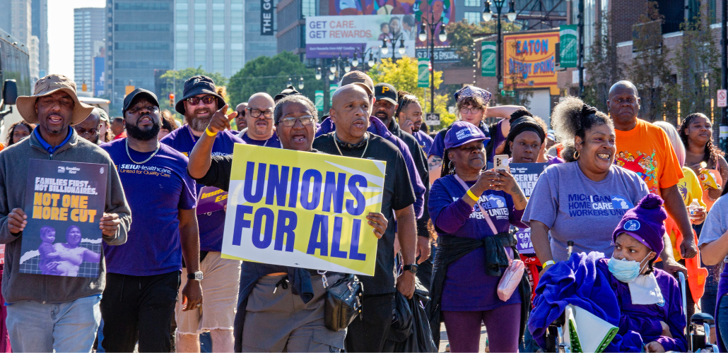 Workers march through a Detroit street, part of a Labor Day march. The people visible are mostly wearing purple SEIU T-shirts, many are Black and one holds a big printed sign readgin "Unions for all" on a yellow background. Another printed sign says "Families first, not billionaires. Not one more cut." One woman in a "Michigan home care workers" shirt pushes another in a wheelchair, also in purple gear.