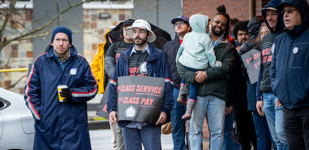 A racially diverse, young, mostly male crowd stands in front of a post office. Some people wear postal gear, others wear "Fight Like Hell" logo T-shirts, and some hold printed "First class service, first class pay" signs. One man holds a toddler in his arms. People wear coats and hats, and one open umbrella is visible in the back.
