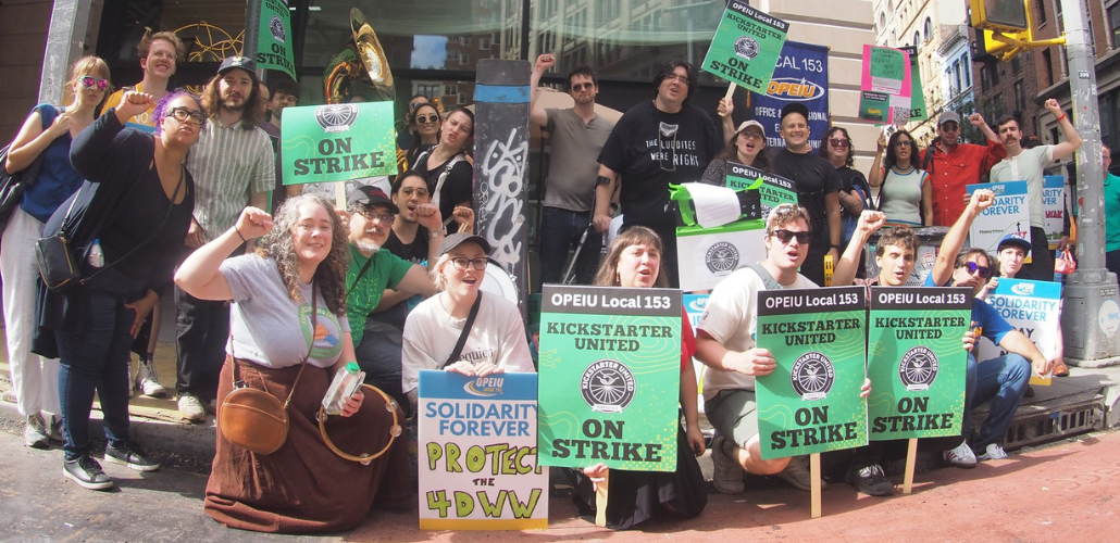 Youngish workers pose with raised fists on a city street. Printed green signs say "OPEIU Local 153, Kickstarter United, On strike," One handwritten sign says "Protect the 4DWW"