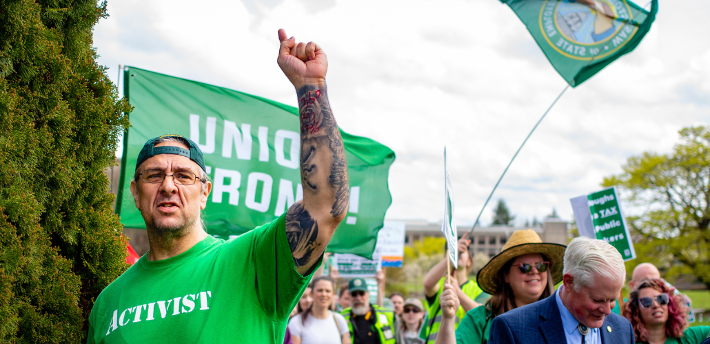 A man in a backwards green ballcap and an AFSCME green T-shirt printed with the word "Activist" raises his heavily tattooed arm up to the sky. Behind him other marchers are visible, crossing the capitol campus, some with signs (not readable) and one waving a Washington state flag on a tall pole.