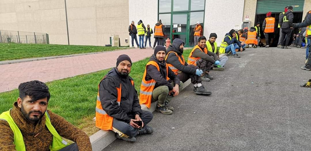 Workers in Italy sitting outside a warehouse on strike in orange vests.