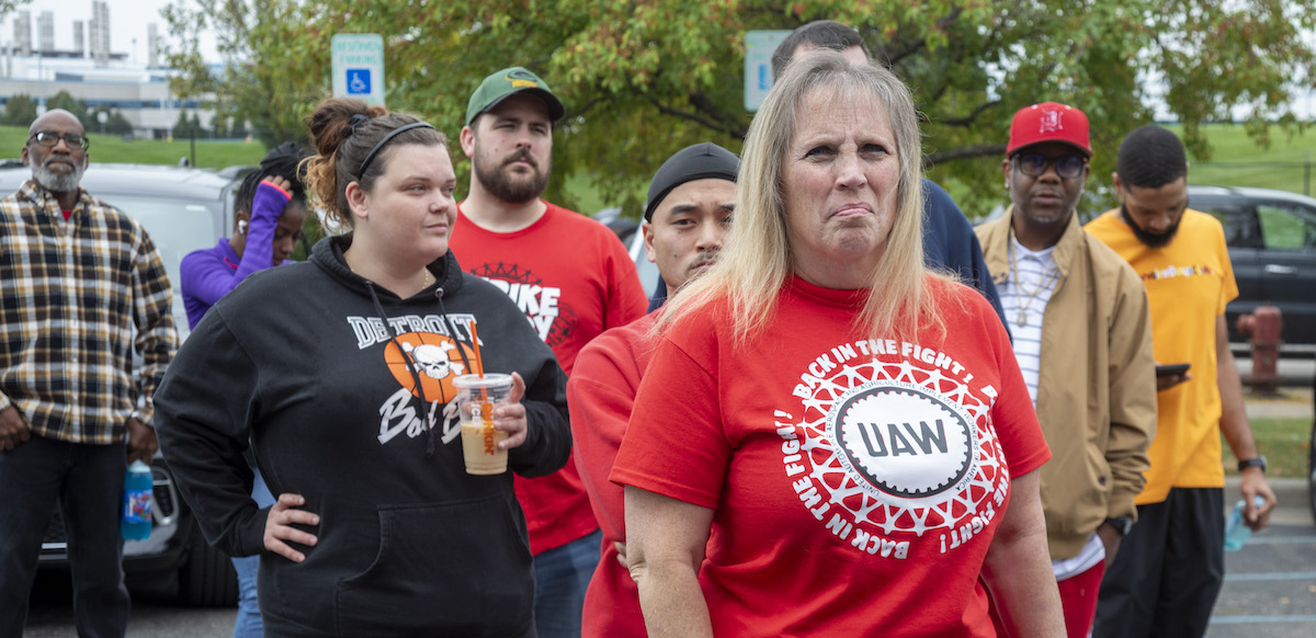 A group of autoworkers stand around outside looking at the camera--the closest is a blonde woman with a red shirt that says “UAW Back in the Fight.”