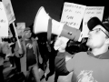 A man yells into a bullhorn with people in the background holding signs--is it blurry and black and white