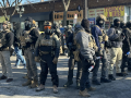 Agents covered head to toe in tactical gear and warm clothing stand in a diamond formation, facing outward, in a city street. Their faces are covered. The vests say "police" though they are not in uniform. They all wear heavy boots and some carry spray cans. Visible behind them is a one-story brick building lined with retail shops, with faded signs reading "8 Street Nourish" and "Tobacco & Mas."