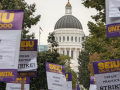 Printed SEIU Local 1000 picket signs reading "Respect state workers!!" and "Unfair practice strike!" are raised aloft with a capitol rotunda visible behind them.