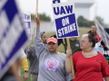 The camera focuses on two women in a crowd of picketers, diverse in race and gender, carrying printed blue and white "UAW on strike" picket signs. The women are wearing different UAW T-shirts. One has the collar cut off, revealing part of a tattoo that reads, in cursive, "darkness light shall prevail." Both women wear serious, determined expressions.