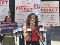 A woman speaks into a microphone outside while others stand behind her with signs that say “Picket ICE”
