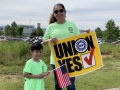 Woman holding "UNION YES" sign and child holding U.S. flag, with VW plant behind