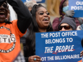 Two Black workers foregrounded in a dense crowd. Man on left wears orange New York Metro APWU T-shirt and lifts his hands to clap. Woman on right has thick eyelashes and holds a blue printed sign: "The Post Office belongs to the people, not the billionaires." Both smiling, energetic, probably chanting.