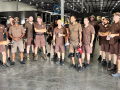 Fifteen workers in brown UPS uniforms stand in a semicircle while another uniformed worker speaks to them