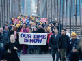 A dense and diverse crowd of people, some wearing yellow hats, marches across the Brooklyn Bridge in early-morning light. At the front are Zohran Mamdani and others carrying a banner that says in big purple letters "Our Time Is Now"