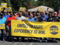 A large group stands behind a yellow banner saying “United to Fight, Experience to Win”
