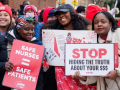 Three Black women nurses pose for camera; two are smiling, one looks determined. Two hold printed NYSNA signs: "Safe nurses = safe patients" and "STOP hiding the truth about your $$$" and the other holds a handwritten sign, partly covered, long text. Lots of red NYSNA hats are visible in the dense, upbeat crowd behind them.