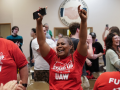 A Black woman in a red "Stand up UAW" T-shirt throws her hands in the air in celebration, phone in one hand, tears in her eyes. Other workers in matching shirts, and other people, stand nearby, all smiling, some clapping. In a room, looks like a union hall, with a logo on the wall that says "United we stand, divided we fall" around a handshake. 