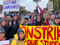 Smiling strikers pose outdoors with a bright yellow banner handpainted "On strike for our schools" in red. Most people visible in this photo appear to be white or Asian women. Many wear UESF hats. Other visible handmade signs say "Boo Su, give us a fair contract now!" "Keep our counselors" and "Invest in oiur largest asset: children." Palm trees are visible against gray sky. Some people raise fists or a victory/peace sign.