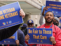 People (mainly Black men are visible) outside hold printed AFGE signs reading "Proud veterans serving veterans," "Be fair to those that care," and "VA workers deserve dignity, fairness, and respect."