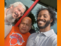 Three workers pose for a group selfie in front of the store. They read as three men of different races. The man in the middle is wearing an orange Knicks T-shirt.