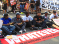 Fast food strikers, mostly young and Black, a mix of genders, sit in an intersection, linking arms. A long red banner laid out in front of them says "Whatever it takes." In the crowd behind them, some people hold a white handlettered banner: "Give us this day our daily bread."