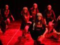 Seven women in long skirts and kerchiefs kneel, their arms in motion, apparently mid-dance. They are on a stage, bathed in red light.