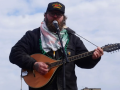 Emmett Doyle, a bearded white man wearing a "Class war veteran" hat, a keffiyeh, and a Carhartt jacket, plays an Irish bouzouki. He is outdoors, silhouetted against a blue sky with thin white clouds.